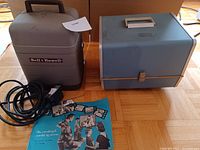 Bell and Howell projector and Argus slide projector with power cords, cases, and a vintage instruction manual on wooden flooring