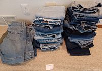Two stacks and one pile of assorted blue denim jeans and shorts on carpeted floor next to a wall outlet. Most pieces are folded, showing significant wear and fraying, representing small ladies sizes.