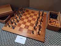 Chess board with metal Greek-style chess pieces set on it and visible wooden storage boxes with additional pieces.