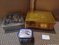 Photo of three decorative metal storage boxes placed on carpet. Includes silver-tone box with Victorian horse-drawn carriage design, gold-tone box with sunburst and floral accents, and small blue J&P Coats sewing thread tin.