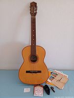 Front view of Giannini 6-string acoustic guitar alongside vintage beginner books, guitar strap, extra strings, and capo arranged on a blue surface.