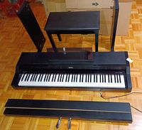 Full view of digital piano with detached pedal board, black bench, and one side panel visible on floor