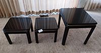 Three black wood tables of varying sizes arranged side by side showing glossy black mirrored finish tops and metallic trim edges.
