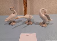 Three white porcelain duck figurines arranged side by side in front of a ruler, showing size and overall condition with visible damage to bill on one duck.