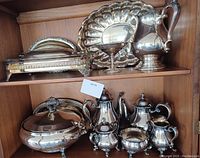 Photo showing a collection of silverplate items including tea and coffee pots, creamers, sugar and waste bowls, trays and platters on a wooden shelf.