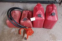 Four red plastic gas cans in varying sizes grouped together with a red manual fuel transfer pump attached to black hoses. Items are on concrete floor showing usage wear.