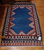 Photo of small area rug with mainly blue central area and red, white, black geometric borders on wooden floor.