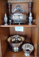 Set of sterling and silverplate salt and pepper shakers, silverplate bowls, trivets, and casserole displayed in a wooden cabinet shelf.