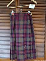 Front view of the magenta and brown plaid wool kilt hanging on a hanger, showing pleats and fabric pattern