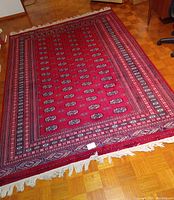 Full view of deep red rectangular Bukhara rug laid flat on parquet floor showing repeating boteh motifs and white fringe ends