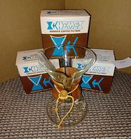Photo of the Chemex coffee maker in front of three separate unopened boxes of Chemex bonded coffee filters, showing hourglass glass shape, wood collar, leather tie, and blue and brown packaging of filters.