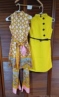 Front view of yellow textured sleeveless dress with black buttons next to colorful patterned sleeveless pants suit with matching belt and pants.
