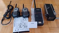 Photo shows two walkie talkies, a Pro-50 police scanner, mini TV, cables, and user manuals on a wood floor.