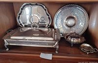 Six silverplate items including two large platters, rectangular covered casserole dish, and covered candy dish displayed on shelf.