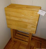 Four blond wood foldable TV trays stacked upright against a wall showing the top surfaces and folded legs.