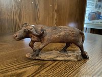 Wooden carving of an African buffalo positioned on a wood surface with a platform base, showing side profile view.