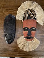Two carved African masks side by side on a wooden surface, one painted with natural fibers and one dark wood carved mask.