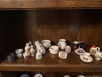 Shelf with assorted UK memorabilia miniatures including jugs, bowls, casks, napkin rings, bulldog salt shaker, shot glass all featuring various UK crests and designs in ceramic and metal under 3 inches.