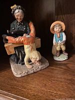 Figurines including elderly woman with child in orange pants and boy in hat and blue vest, displayed in a wooden shelf.