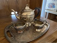 Photo showing a tarnished silver tea set on wooden table including a tray, a teapot with black handle, a creamer, and a sugar bowl.