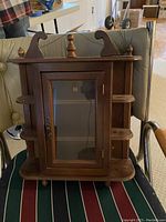 Photo of small wooden wall cabinet with glass front door and two side shelves, decorative top finial and scroll details.