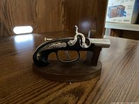 Flintlock pistol lighter on wooden brown base, shown on wooden table with wood panel background.