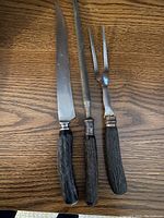 Photo of three carving utensils with dark textured handles: one knife, one fork, and another tool with a long straight handle