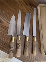Four Japanese Hocho kitchen knives with wooden handles, brass bolsters, and metal blades, arranged on a wooden surface next to the original box.