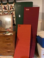Three Therma Rest sleeping pads stacked vertically against wall near wooden furniture and cooler. Sizes and colors visible. Pads are orange, green, and maroon.