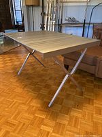 Full side view of aluminum camp table with X-shaped legs and slatted top on parquet floor indoors.