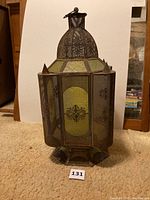 Front view of a tall antique metal candle lantern with decorative glass panels and intricate metal mesh around the sides. The lantern is placed on a beige carpet with a white backdrop.
