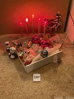 Photo showing lit wooden candle display with red, white, and orange bulbs, small snow-covered tree, and box of assorted ornaments including shiny balls and figurines.