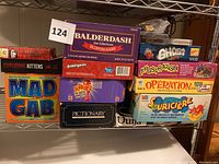 Photo showing many game boxes stacked on metal shelving, titles visible include Balderdash, Scattergories, Exploding Kittens, Mad Gab, Outburst Remix, Pictionary.