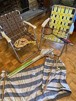 Two folding chairs: one brown and one yellow with woven plastic straps, badminton rackets, rally ball paddle, beach umbrella wrapped, and cotton striped hammock hanging on a metal rack.