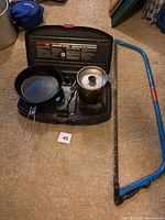 Photo of the propane stove, frying pan, pot with lid inside the stove case, and blue bow saw laid on the floor