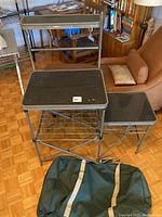 Full view of Coleman folding camp kitchen station with two black top surfaces, wire rack shelf, and green carrying bag beside it on floor.