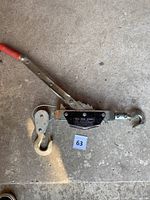 Photo of manual come-along ratchet tool showing the metal body, red handle grip, and two steel hooks.