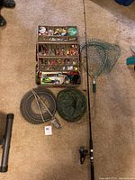 Overview of fishing gear lot including rod and reel, open tackle box filled with assorted lures, a live trap cage, and mosquito net headset pouch.