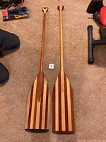 Two wooden paddles laid flat on carpet floor, showing full length with striped wood blades and T-grip handles.