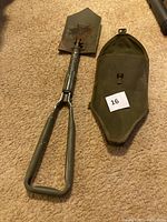 Photo showing the unfolded military-style compact folding shovel with a triangular grip and an olive green metal finish, placed next to the olive green fabric carrying case with a flap and buckle closure.