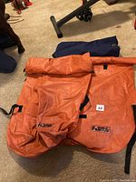 Two large orange waterproof canoe packs with padded shoulder straps, along with a smaller dark blue duffle bag seen from the front.