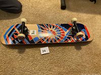 Skateboard bottom side showing colorful geometric design with starburst pattern, white wheels, silver trucks on carpeted floor alongside lot number card 24.