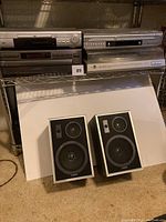 Photo showing two DVD players on metal shelving and a pair of Aiko speakers on the floor beneath the shelf