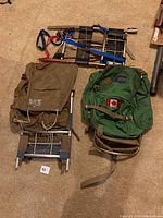 Three external frame backpacks and one external metal frame shown on carpeted floor. Green JanSport backpack with Canadian flag patch, brown canvas backpack, and black metal frame with blue straps visible.