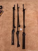Three black collapsible microphone stands lying on carpet floor, showing tripod legs and height adjustment mechanisms.