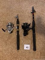 Two telescopic fishing rods with attached spinning reels placed on carpet floor with lot number card