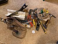 Overview of assorted hand tools including saw, hammer, screwdrivers, pliers, tape measure, utility knife, hand planer, scissors, and small metal scraper, displayed on carpet.