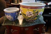 Two Asian flower pots side by side on a green marble-topped chest, showing large yellow pot with floral and bird design and smaller blue pot with floral pattern.