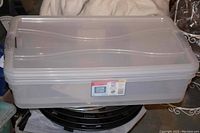 Photo of one large slim clear plastic storage tote with lid stacked on another item; shows overall shape and size.