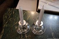 Two depression glass candle stick holders with silver overlay floral patterns around the base and top collar, shown on a green marble surface with white candles inserted.